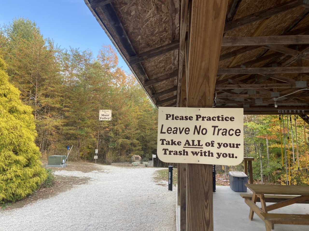 A gravel trail and a covered picnic area in a forested park. A sign reads, "Leave No Trace" at the picnic spot. 